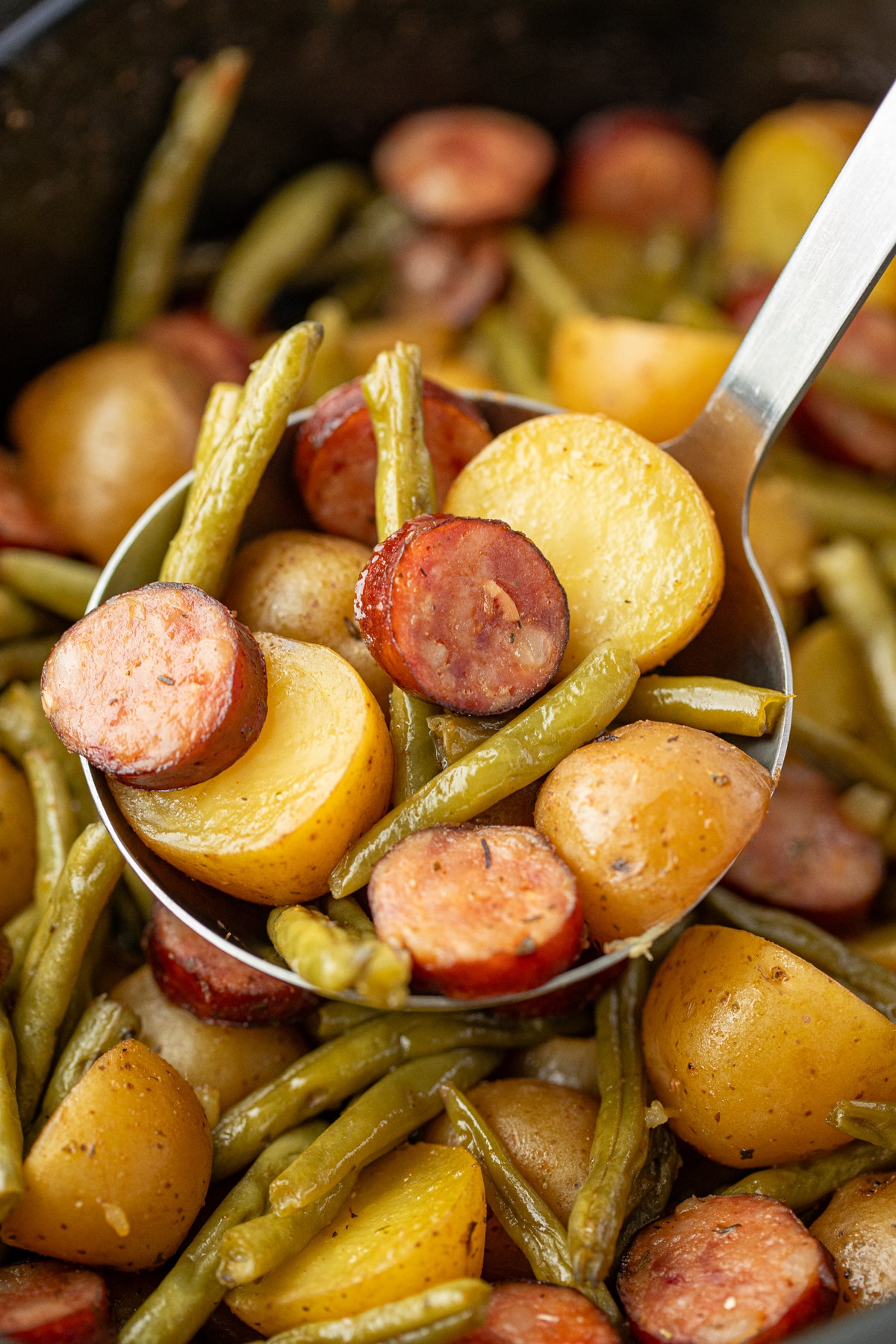 A ladle scooping potatoes, sausage, and green beans from a slow cooker.