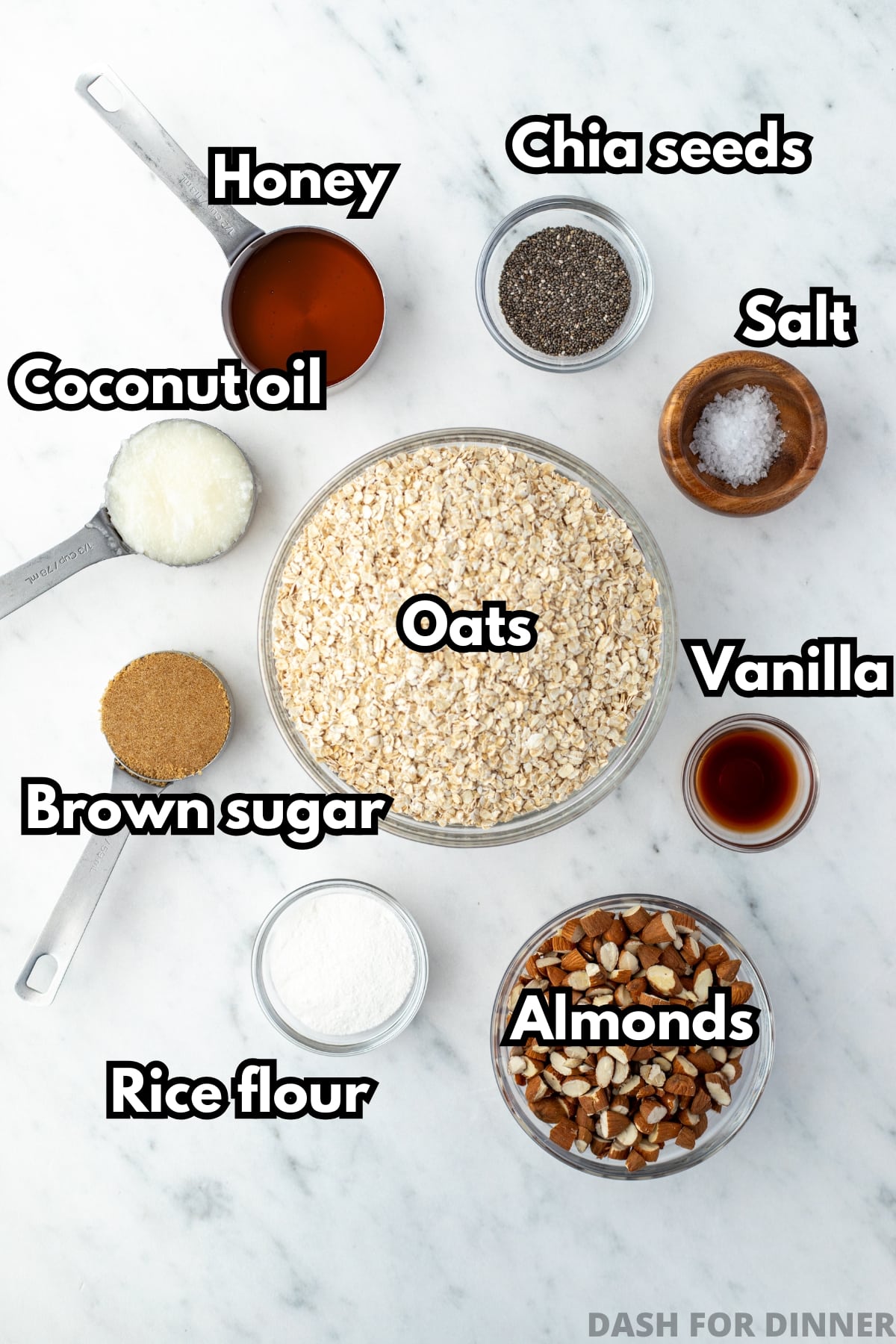 A bowl of quick oats, surrounded by the ingredients needed to make granola, including honey, brown sugar, almonds, vanilla, and chia seeds.