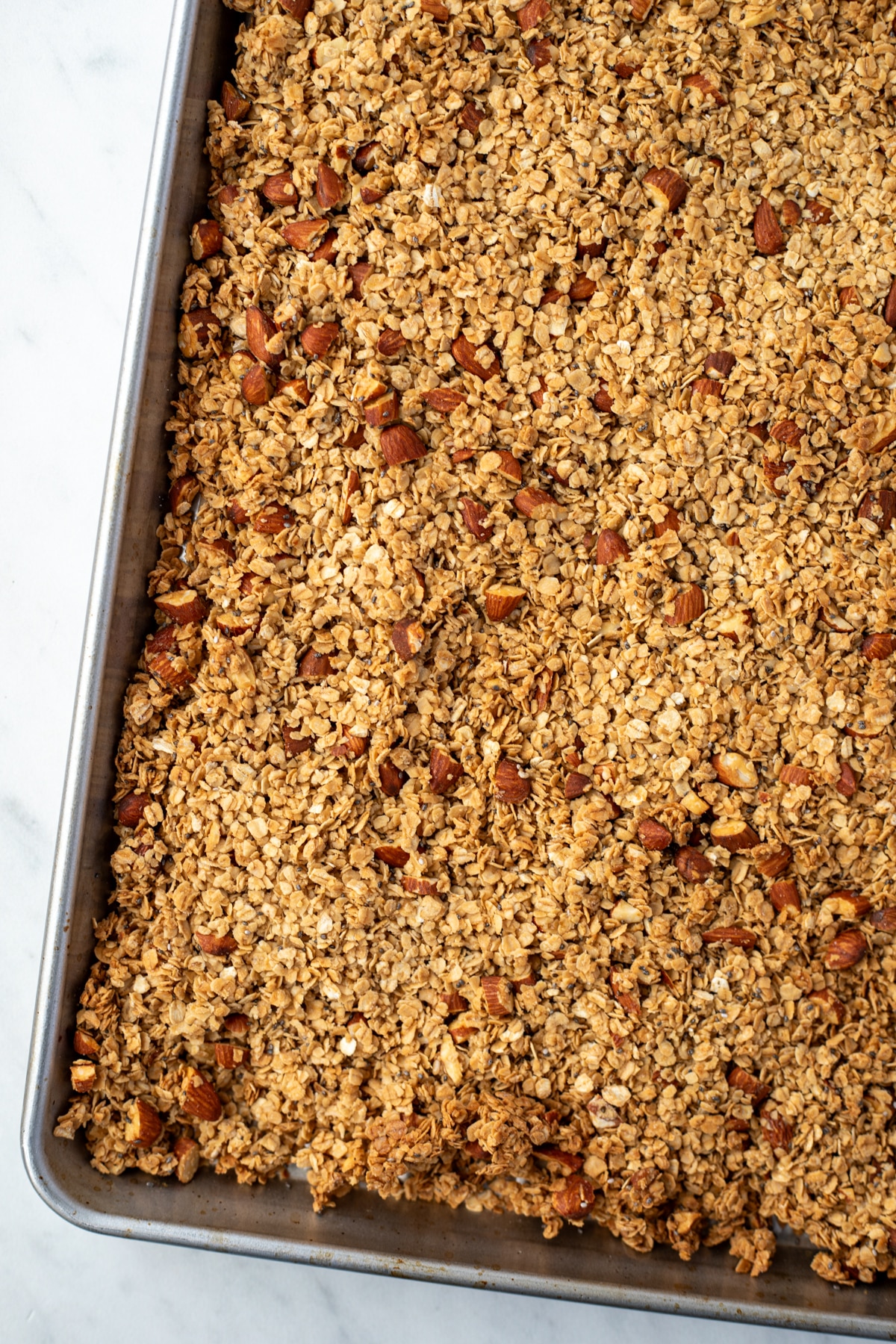 A sheet pan filled with baked granola, dotted with chopped almonds and with lightly browned edges.