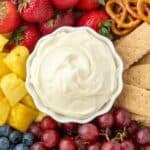 A small bowl of cream cheese dip in the middle of a platter of fruit, graham crackers, and pretzels.
