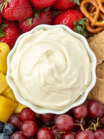 A small bowl of cream cheese dip in the middle of a platter of fruit, graham crackers, and pretzels.