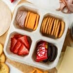 A lunch container with four sections, filled with meat, cheese, crackers, fruit, and a mini chocolate bar.