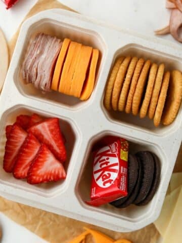 A lunch container with four sections, filled with meat, cheese, crackers, fruit, and a mini chocolate bar.