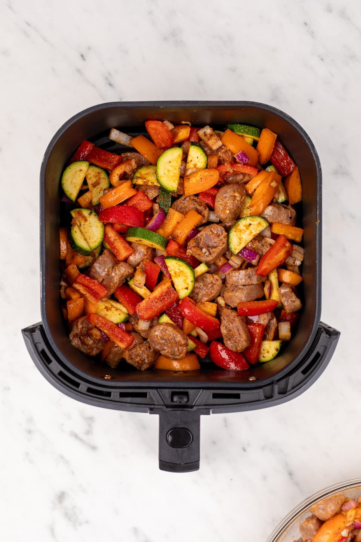 An air fryer basket filled with chicken sausage and vegetables, ready to cook.