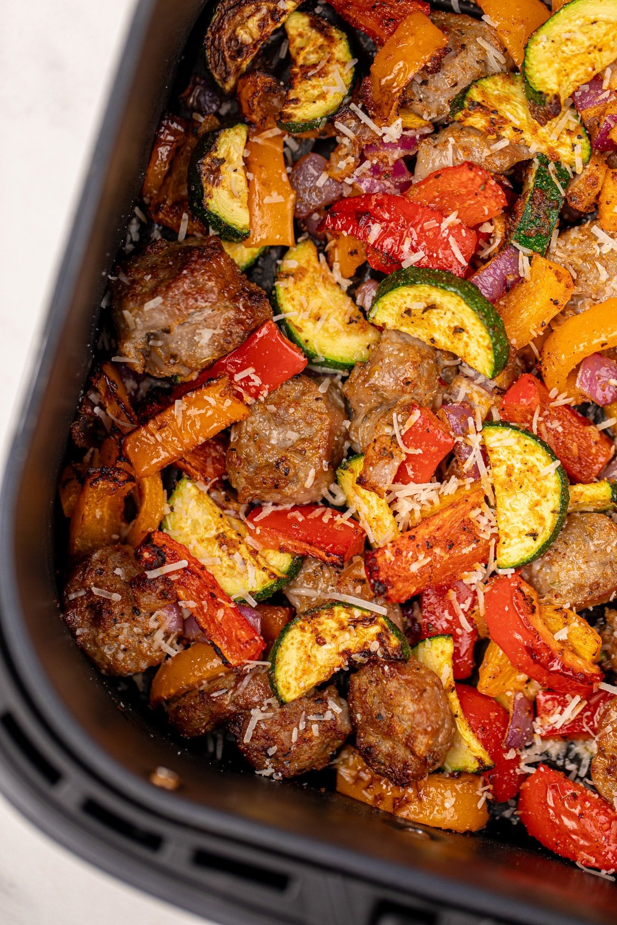 An air fryer basket filled with cooked vegetables, including zucchini, red pepper, and red onion.