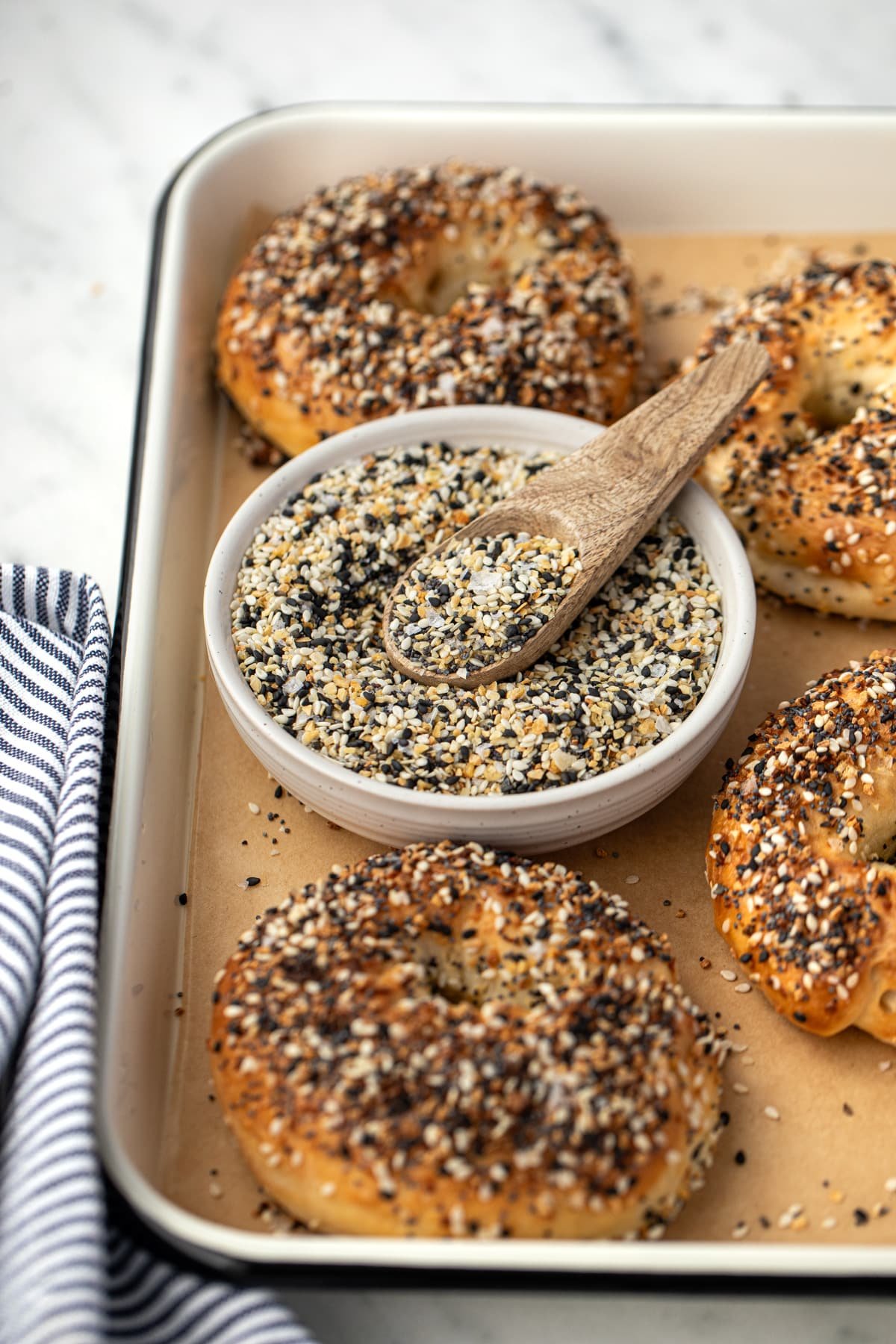A small bowl of everything bagel seasoning on a baking tray surrounded by everything bagels.