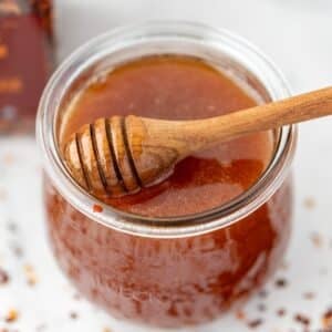 A jar of honey with a loaded honey dipper, and scattered crushed red pepper flakes on the counter.