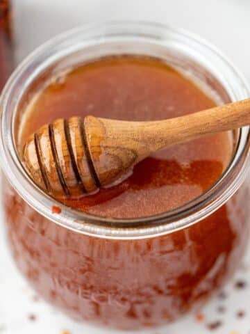 A jar of honey with a loaded honey dipper, and scattered crushed red pepper flakes on the counter.