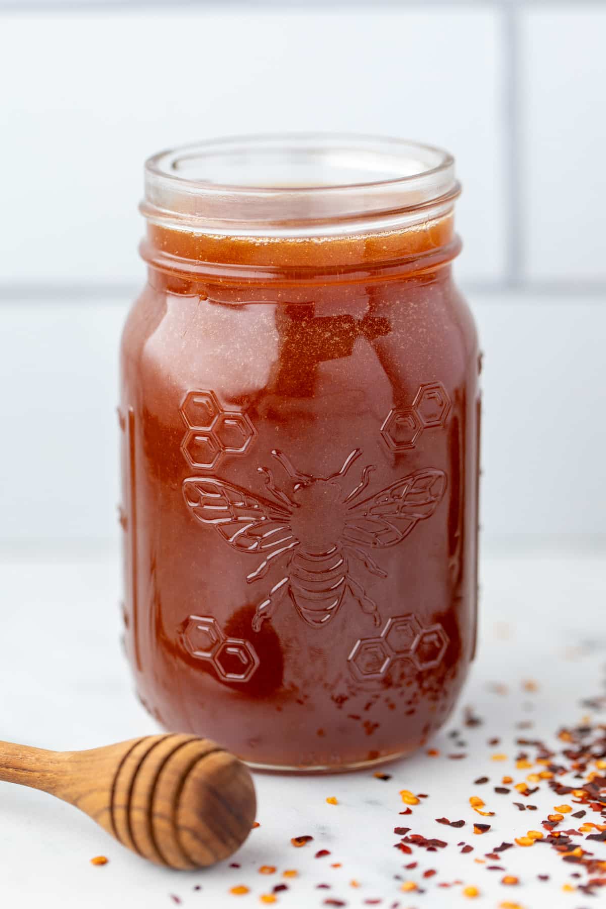 A jar of honey with a honey dipper on the side and crushed chiles on the counter.