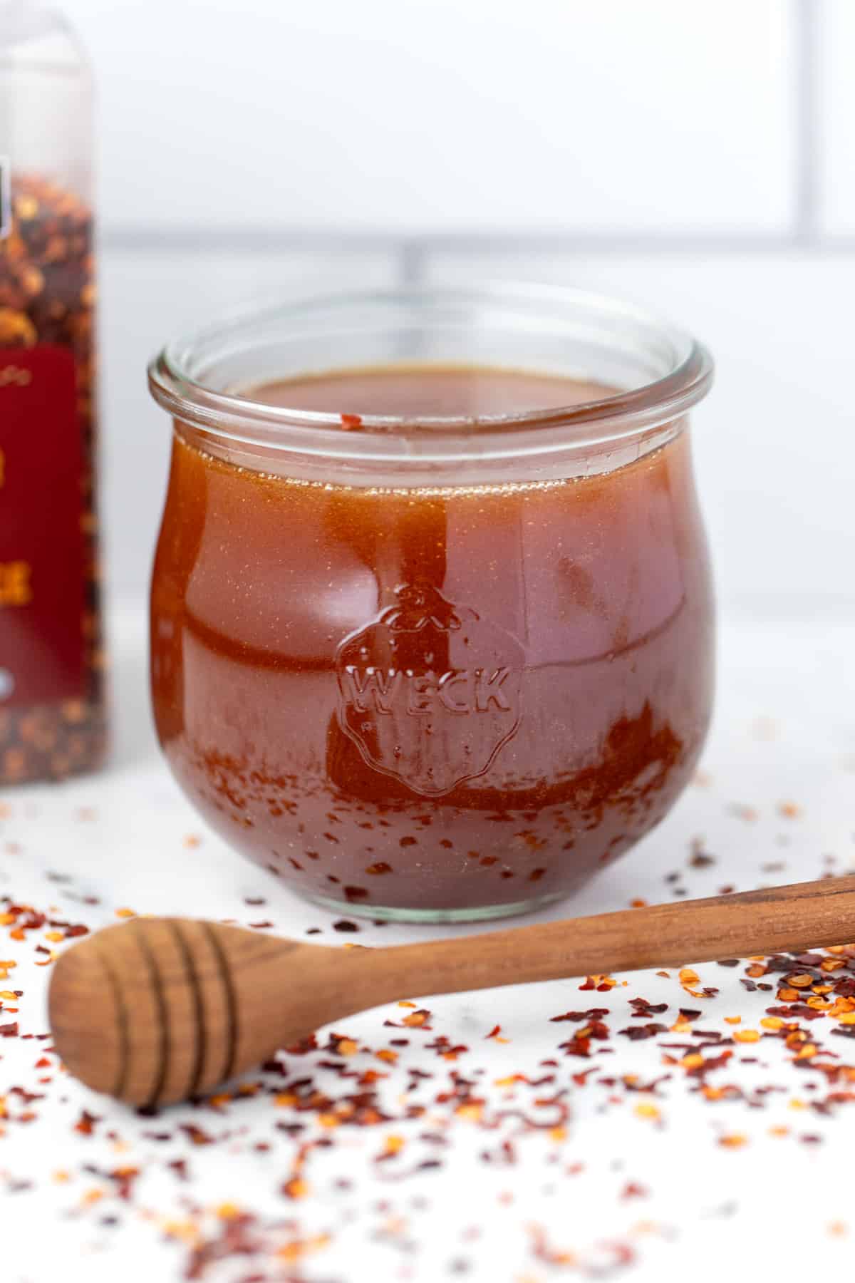 A weck jar filled with honey, with a honey dipper on the counter, along with crushed red pepper.