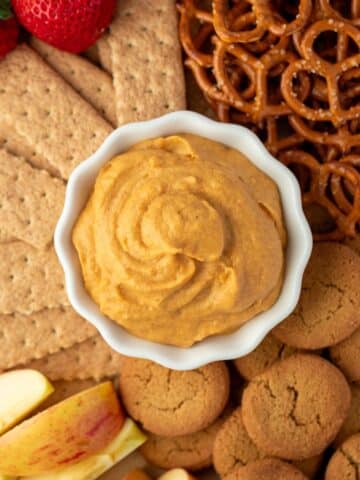 A small bowl of pumpkin dip surrounded by graham crackers, pretzels, ginger snaps, apples, etc.
