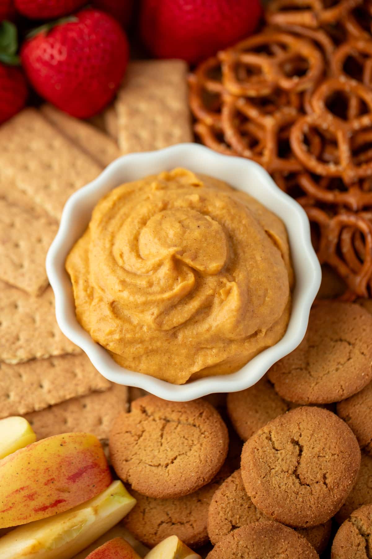 A small bowl filled with pumpkin dip, surrounded by graham crackers, ginger snaps, pretzels, and fruit.