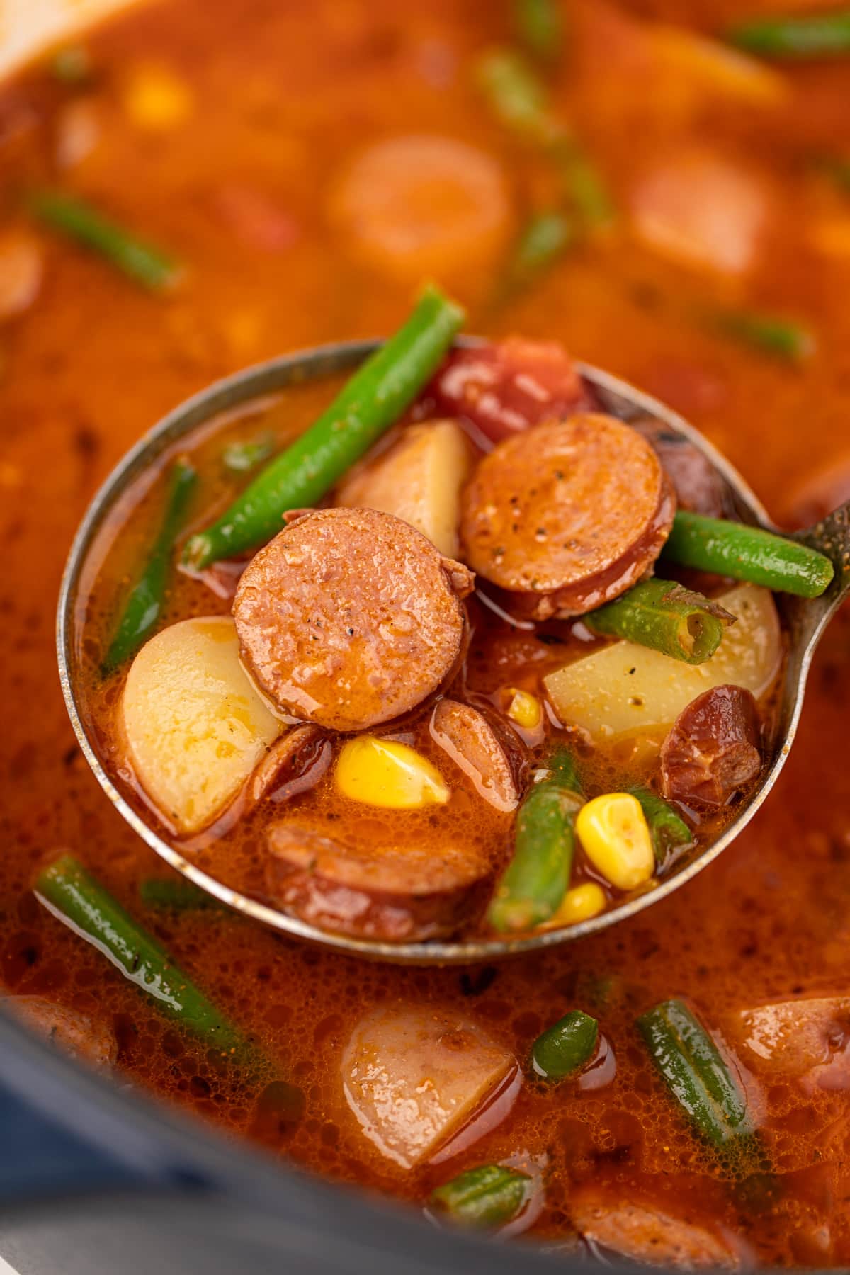 A ladleful of soup that includes potatoes, sausage, green beans, and corn.