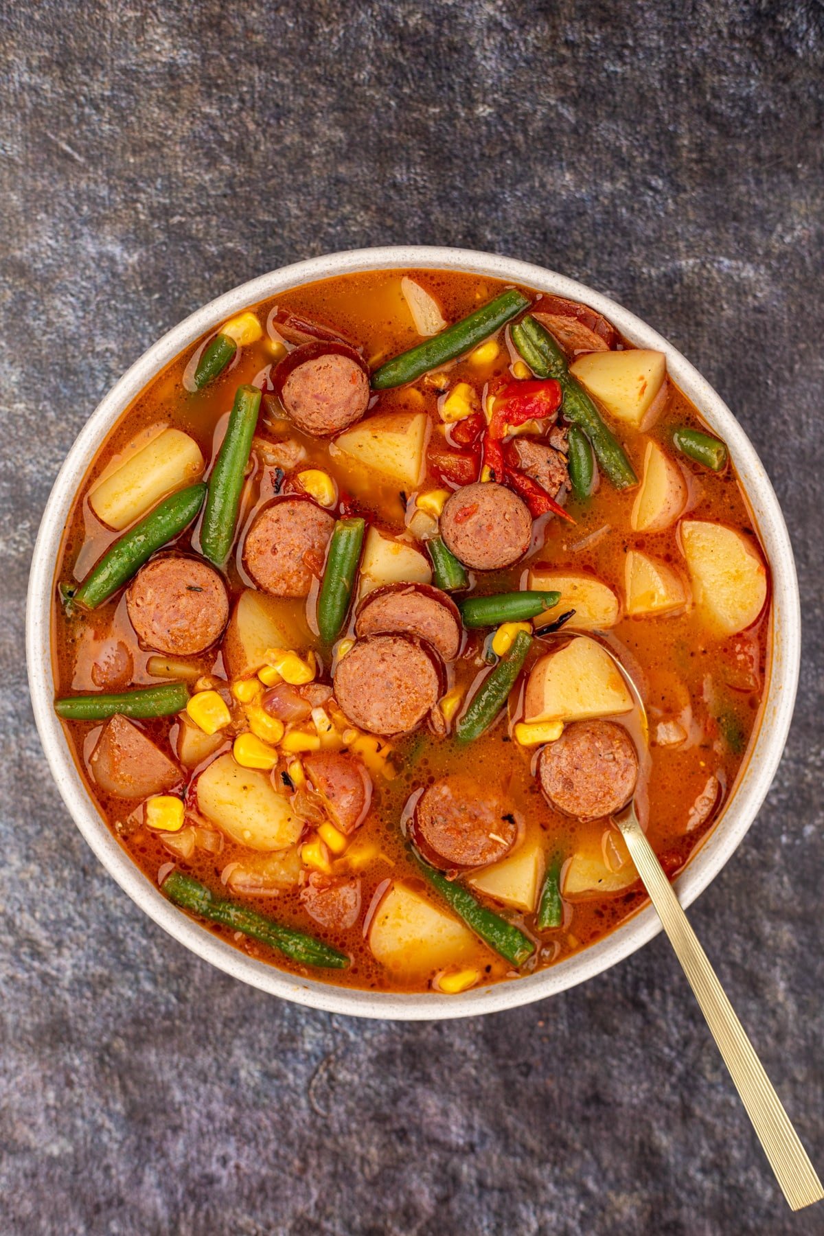 A bowl of soup with chunks of potato, smoked sausage, green beans, and corn.