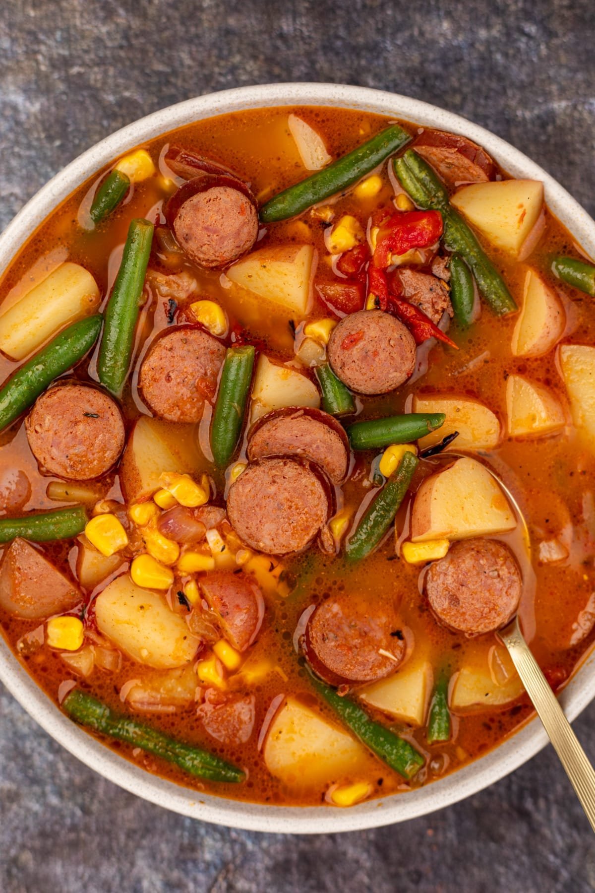 A bowl of hearty stew featuring sliced sausage, potatoes, green beans, and corn.
