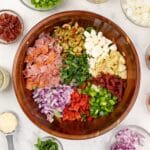 A wooden bowl featuring a variety of salad toppings, including red onion, olives, sundried tomatoes, etc.