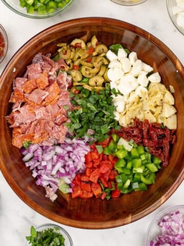 A wooden bowl featuring a variety of salad toppings, including red onion, olives, sundried tomatoes, etc.
