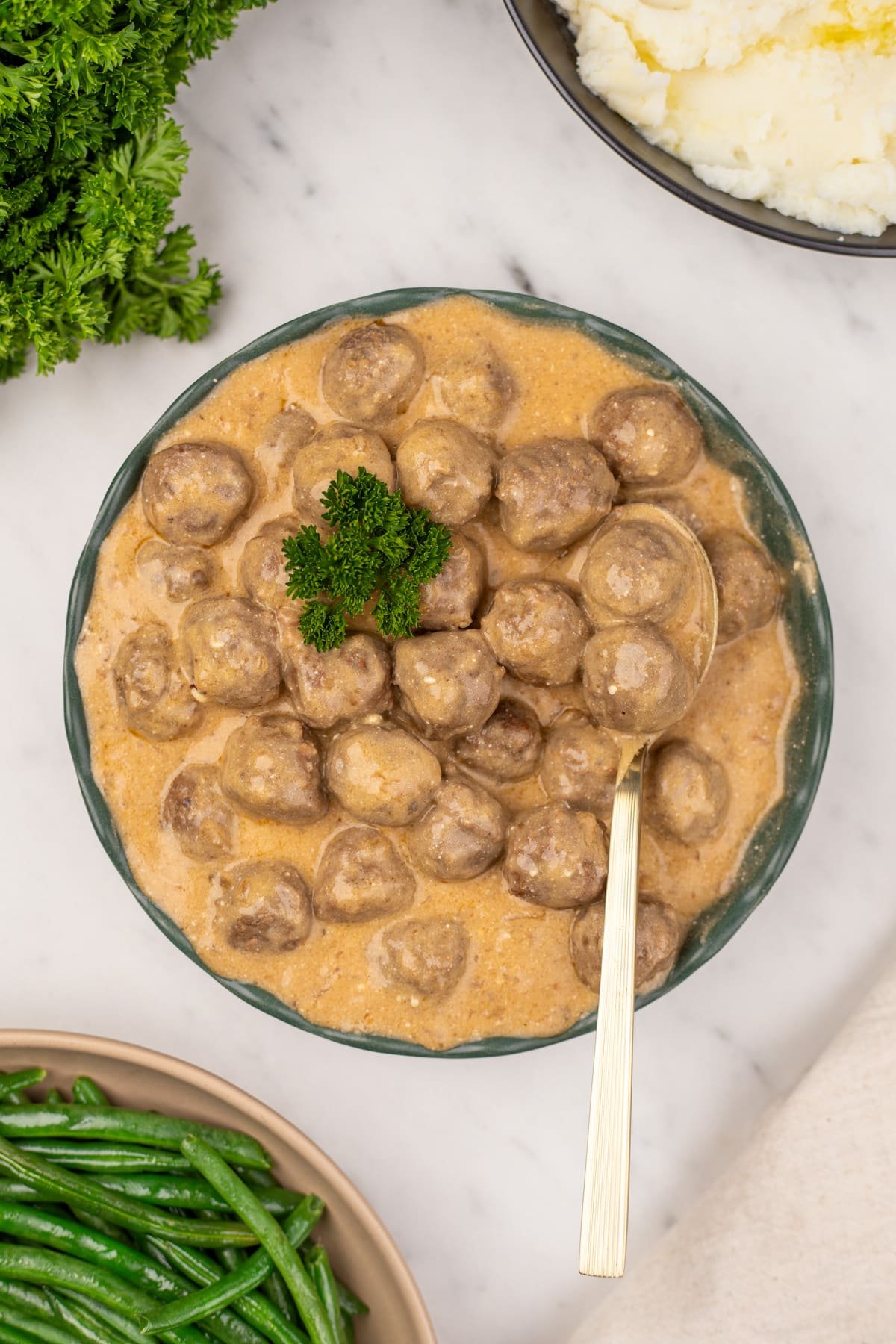 A bowl of creamy meatballs garnished with parsley, with a serving spoon in the bowl.
