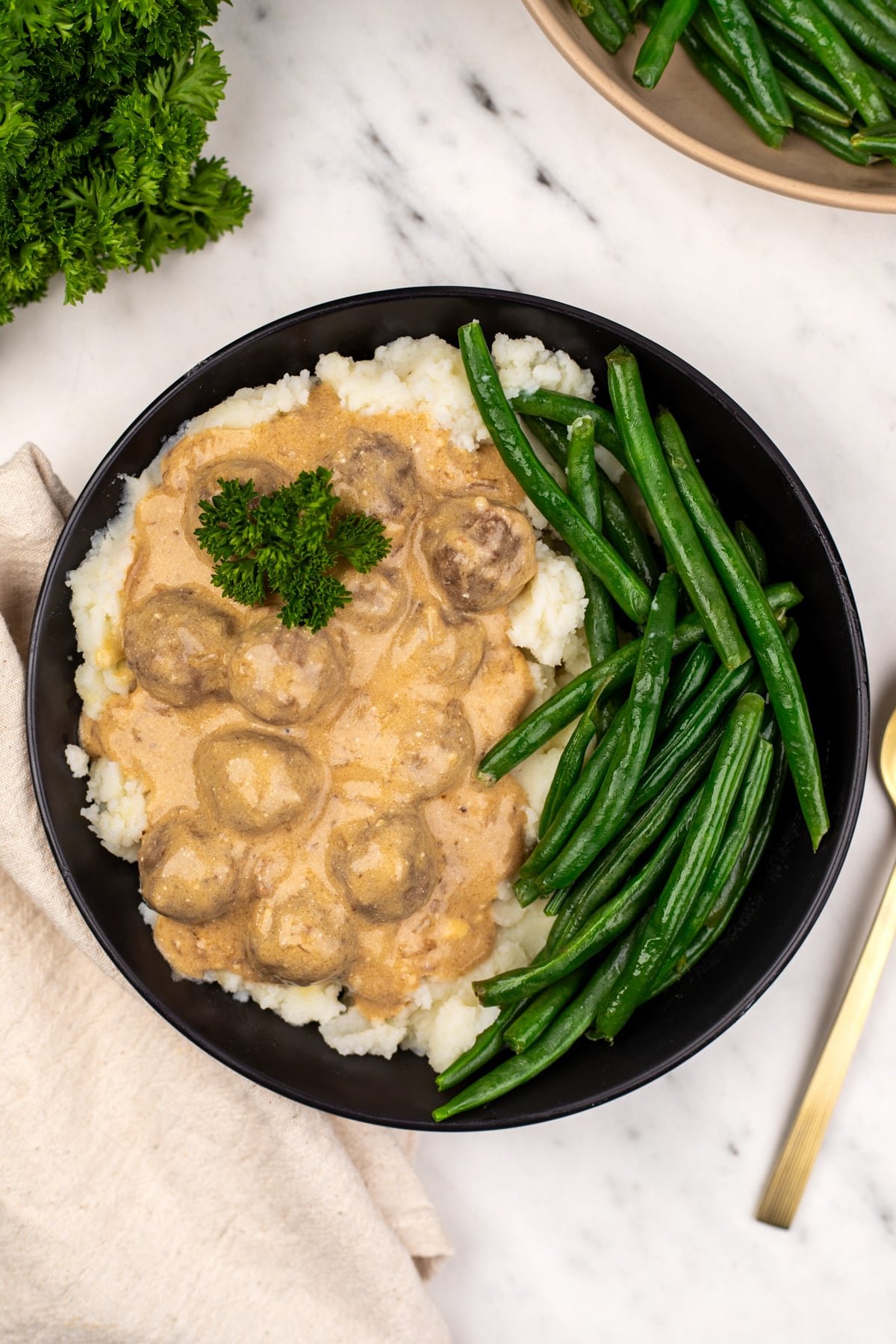 A bowl featuring mashed potatoes, steamed green beans, and swedish meatballs garnished with parsley.