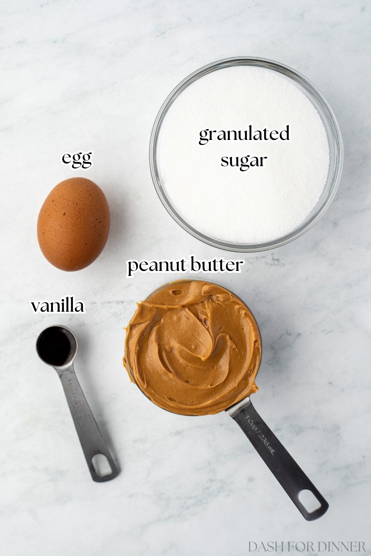 A bowl of sugar, a measuring cup of peanut butter, and egg, and some vanilla extract.