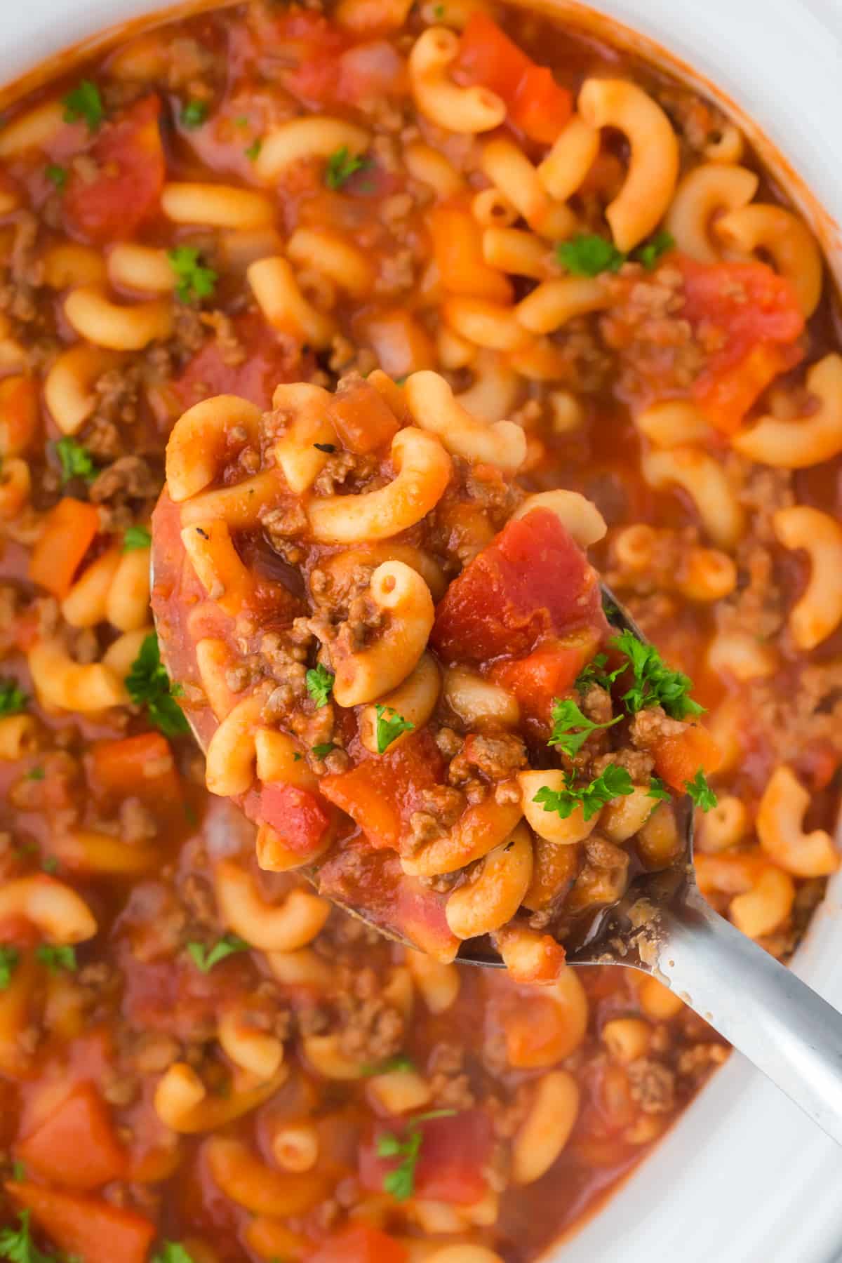 A spoon holding up a large amount of goulash with ground beef and macaroni.