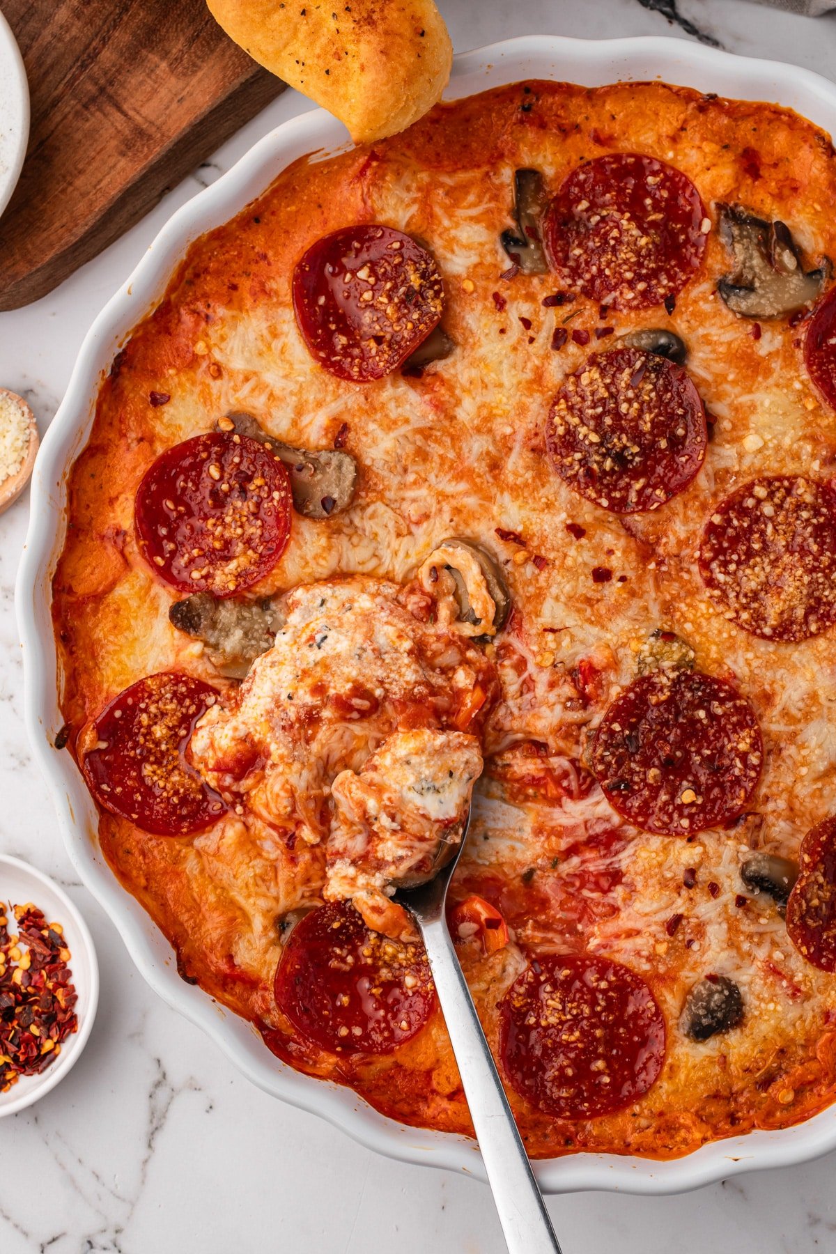 A spoon with melted cheese and cream cheese, inside of a round baking dish filled with pizza dip.