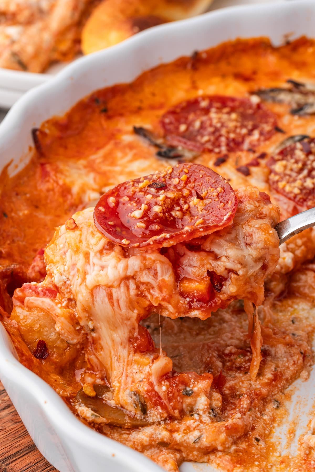 A spoon lifting gooey cheese dip from a baking dish, topped with pepperoni.