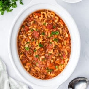 A slow cooker filled with goulash, containing ground beef, elbow macaroni, and diced tomatoes.