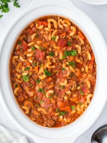 A slow cooker filled with goulash, containing ground beef, elbow macaroni, and diced tomatoes.
