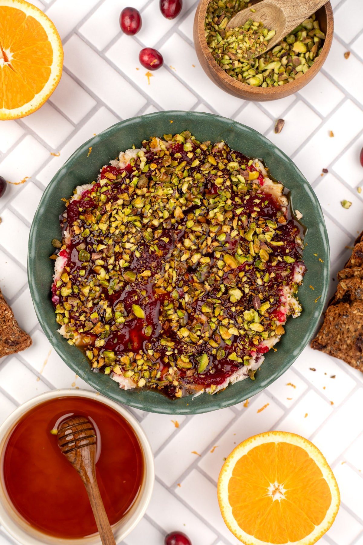 A cranberry and pistachio dip on a white tile background, with a bowl of honey to the left.