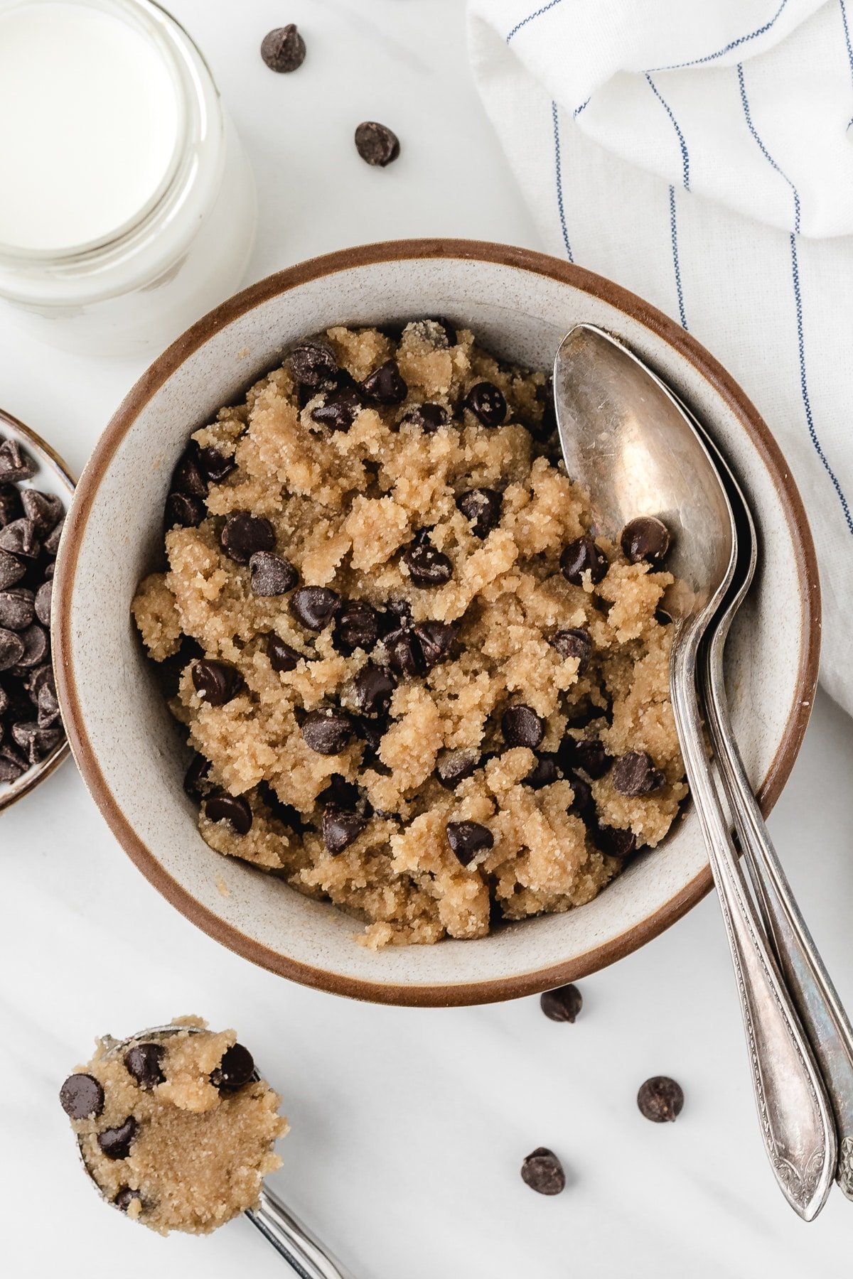 A bowl of chocolate chip cookie dough with a spoon on the side, and a glass of milk just out of view.
