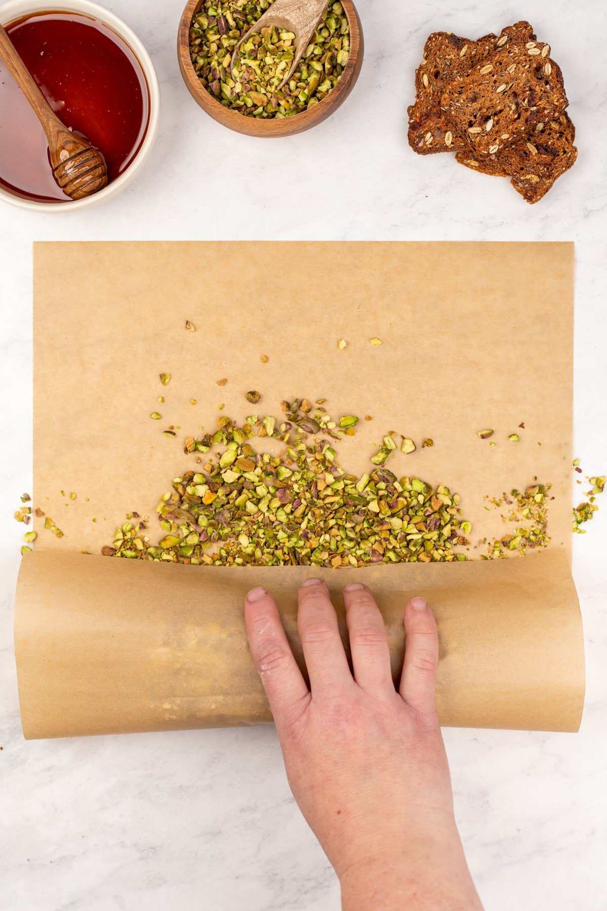 Rolling parchment paper to coat a cheese log in crushed pistachios.