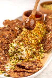 A honey dipper drizzling honey on top of a goat cheese log coated in crushed pistachios.