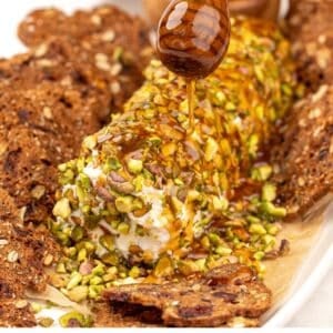A honey stick drizzling honey on a goat cheese log covered in pistachios.