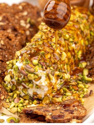 A honey stick drizzling honey on a goat cheese log covered in pistachios.