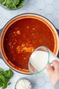 Pouring heavy cream into a dutch oven filled with a tomato based soup.