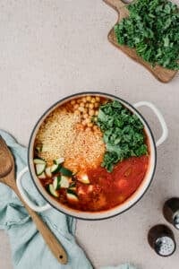 Adding orzo, kale, and vegetables to a Dutch oven filled with soup.