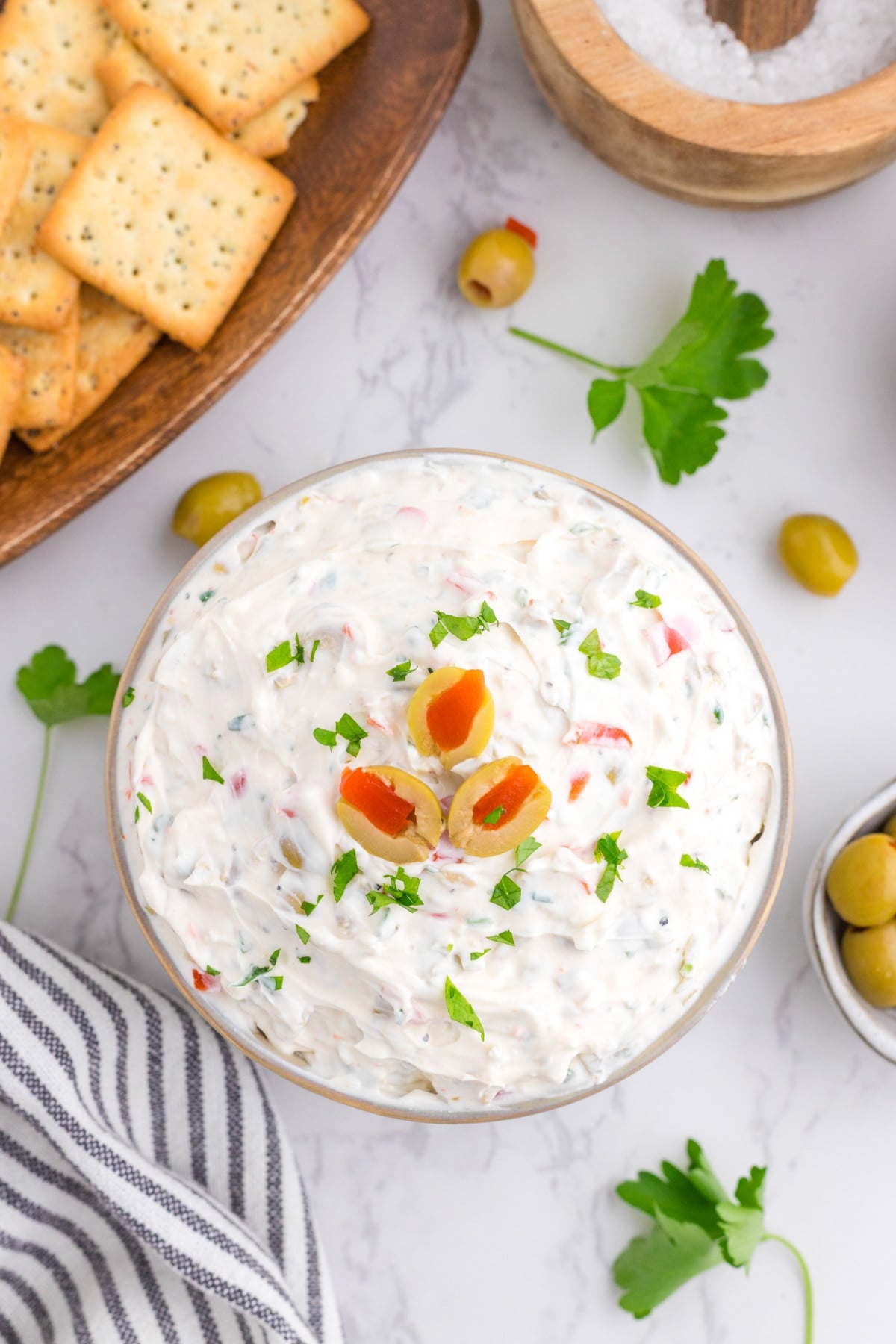 A bowl of olive dip surrounded by crackers, fresh parsley, and a striped napkin.