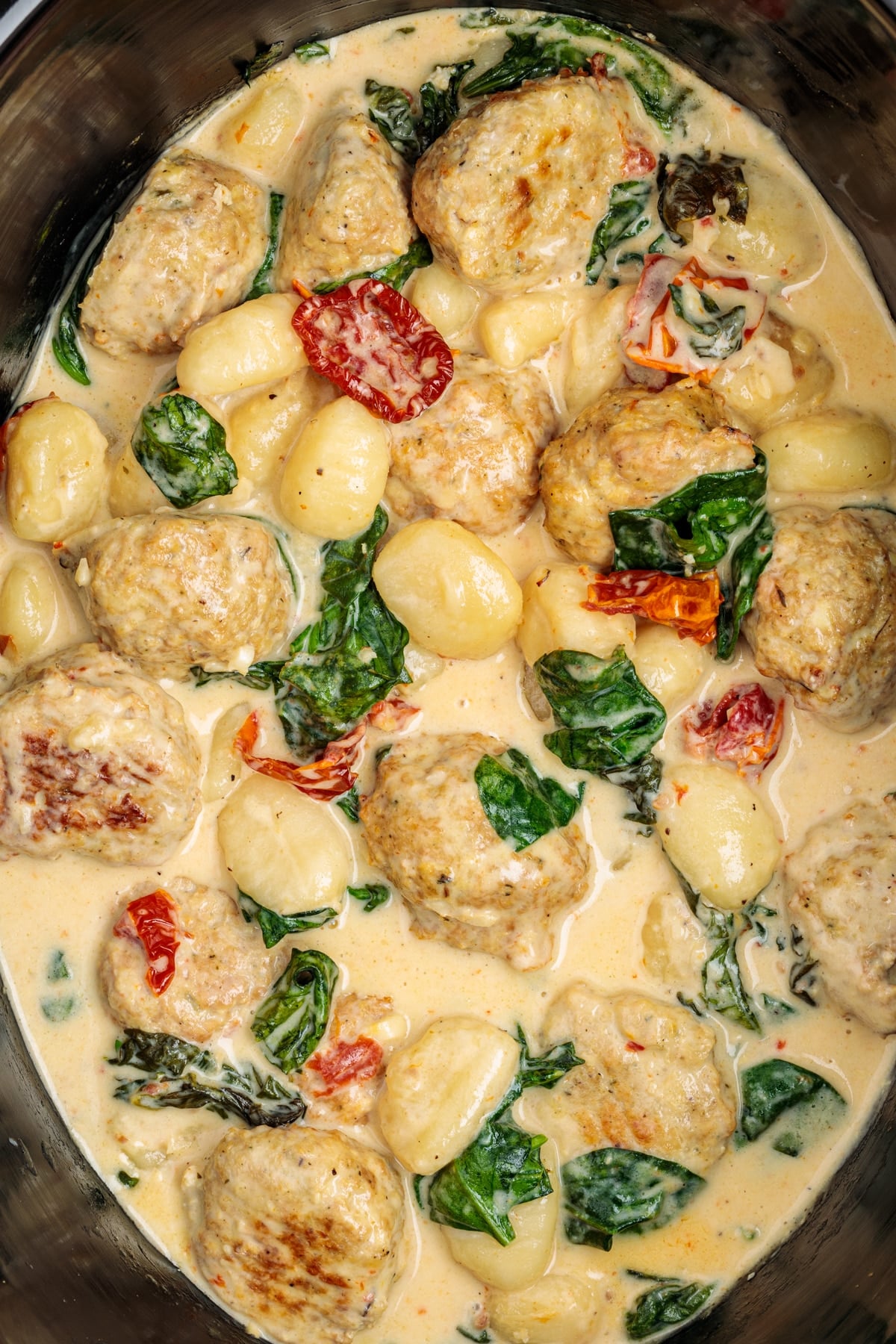 A slow cooker insert filled with a creamy sauce, chicken meatballs, gnocchi, spinach, and sundried tomatoes.