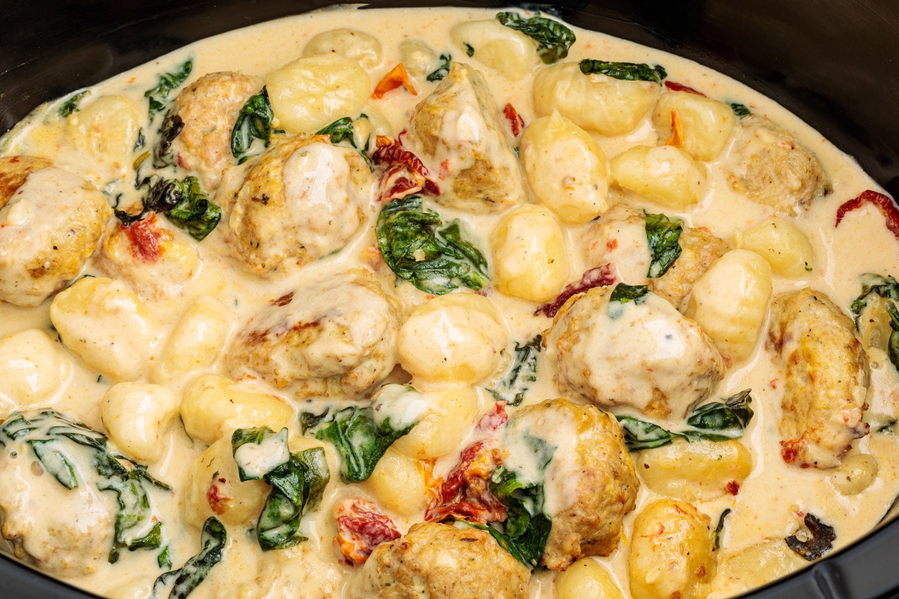 A slow cooker filled with chicken meatballs, gnocchi, spinach, and a creamy sauce.