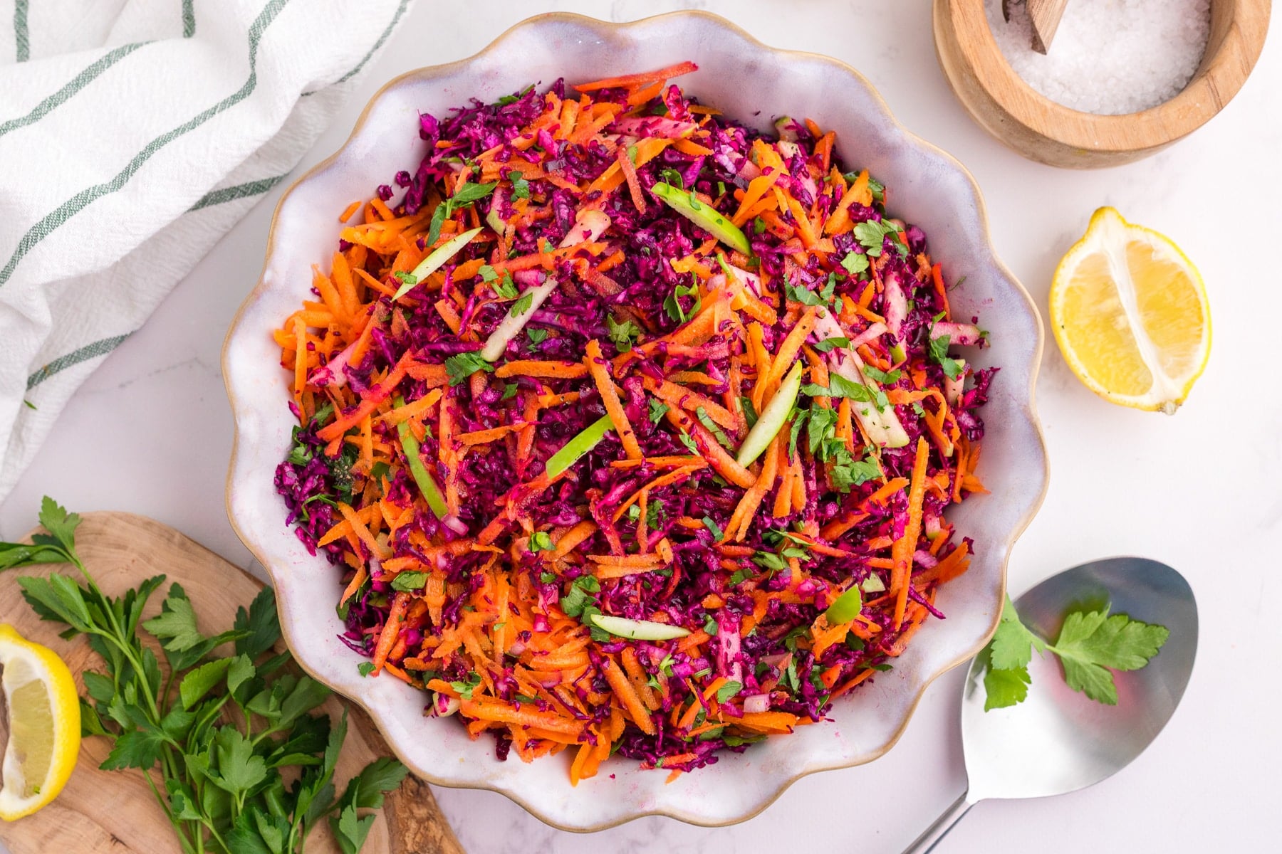 A bowl of cabbage and carrot salad on a white countertop, with lemon and parsley on the table.