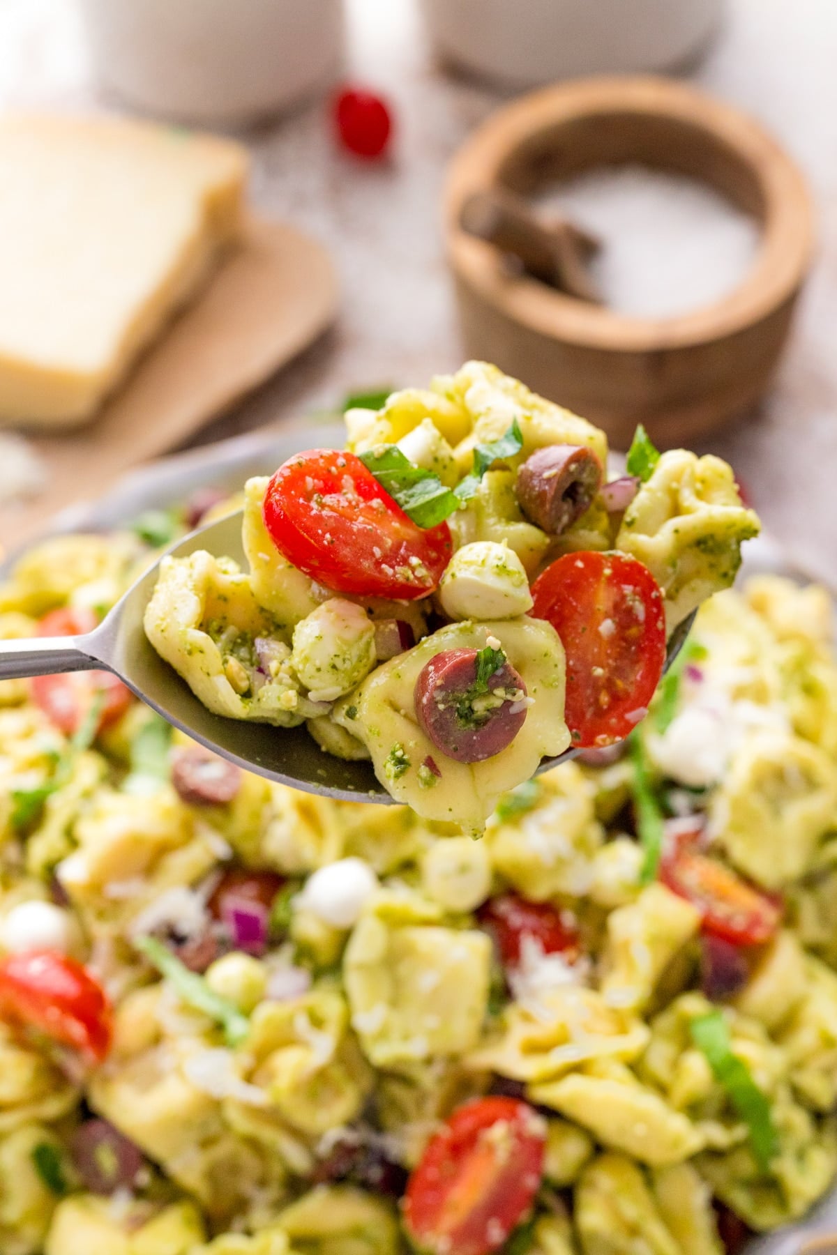 A serving spoon taking a portion of tortellini pasta salad from a large bowl.