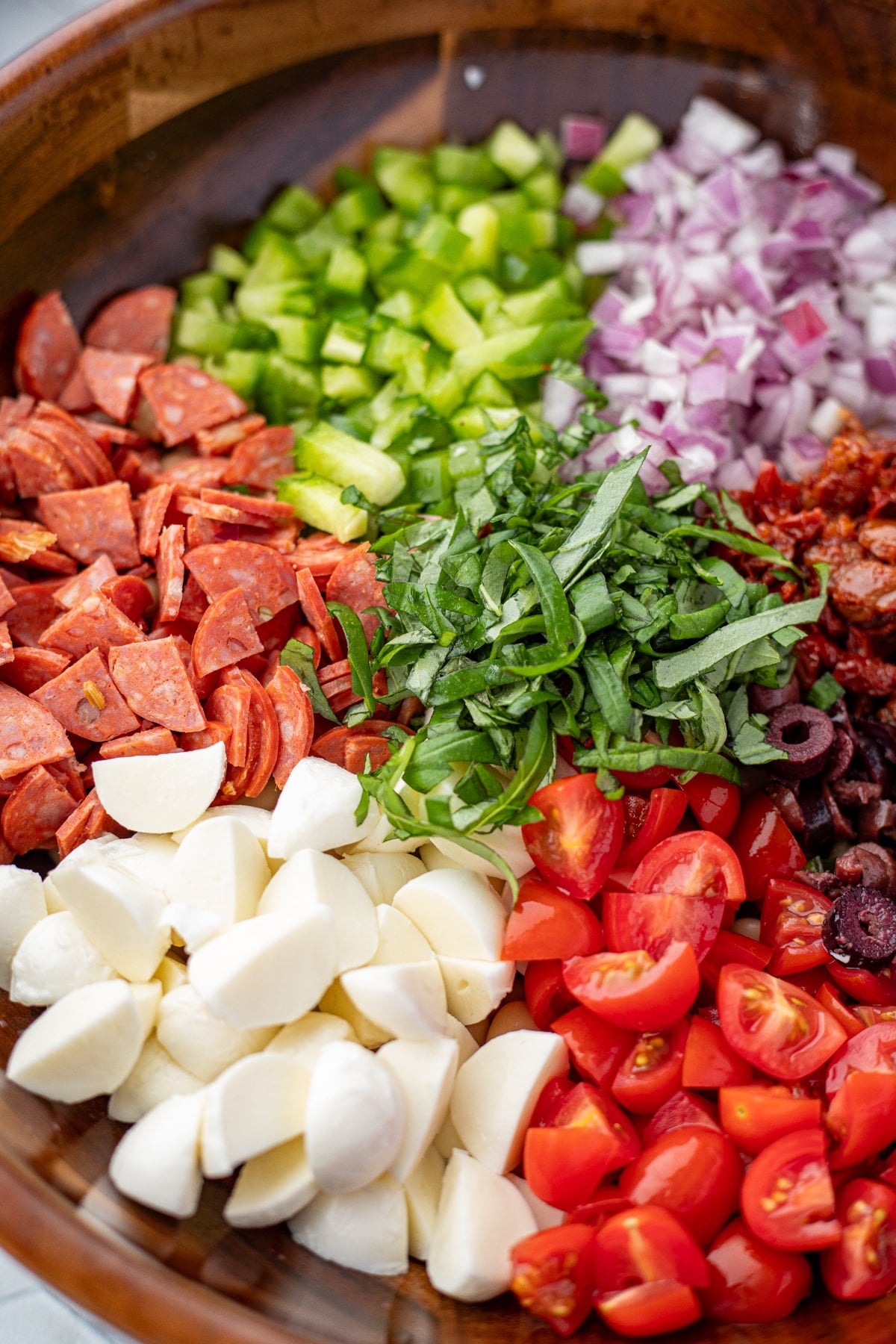 Veggies, pepperoni, mozzarella, and fresh basil arranged in a circular pattern in a wooden bowl.