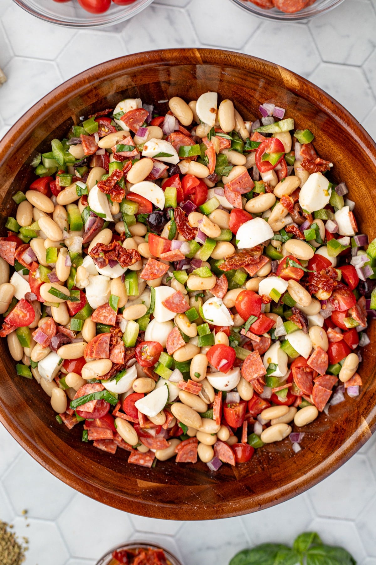 A wooden bowl filled with bean salad, dotted with mozzarella balls, pepperoni, and sundried tomatoes.