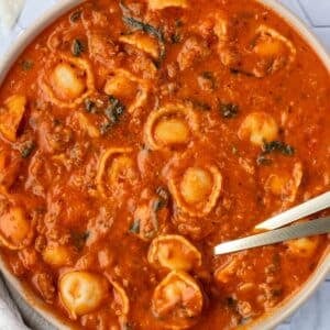 A bowl of soup with a tomato base, filled with tortellini, sausage crumbles, and spinach.