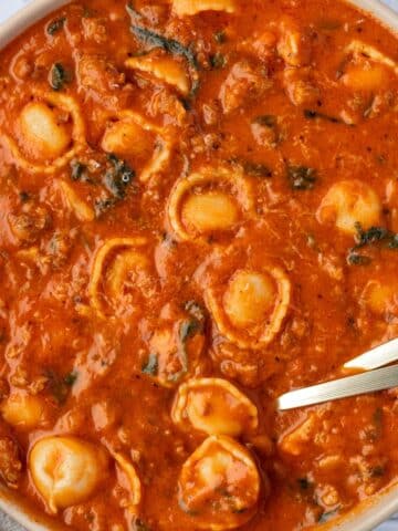 A bowl of soup with a tomato base, filled with tortellini, sausage crumbles, and spinach.