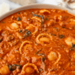 A spoon taking tortellini, tomato, and sausage soup from a serving bowl.