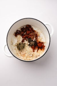 A soup pot with onion, garlic, seasonings, and sun-dried tomatoes.