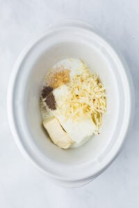 A slow cooker with cream cheese, cheese, seasonings, milk, and butter in the bottom.